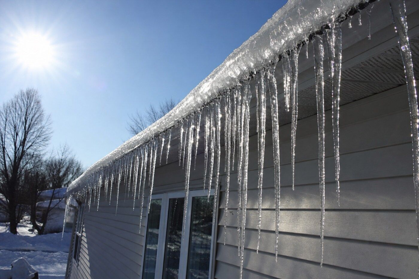 Illustrative image of a thick ridge of ice and long icicles hanging from the eaves of a house in winter sun.